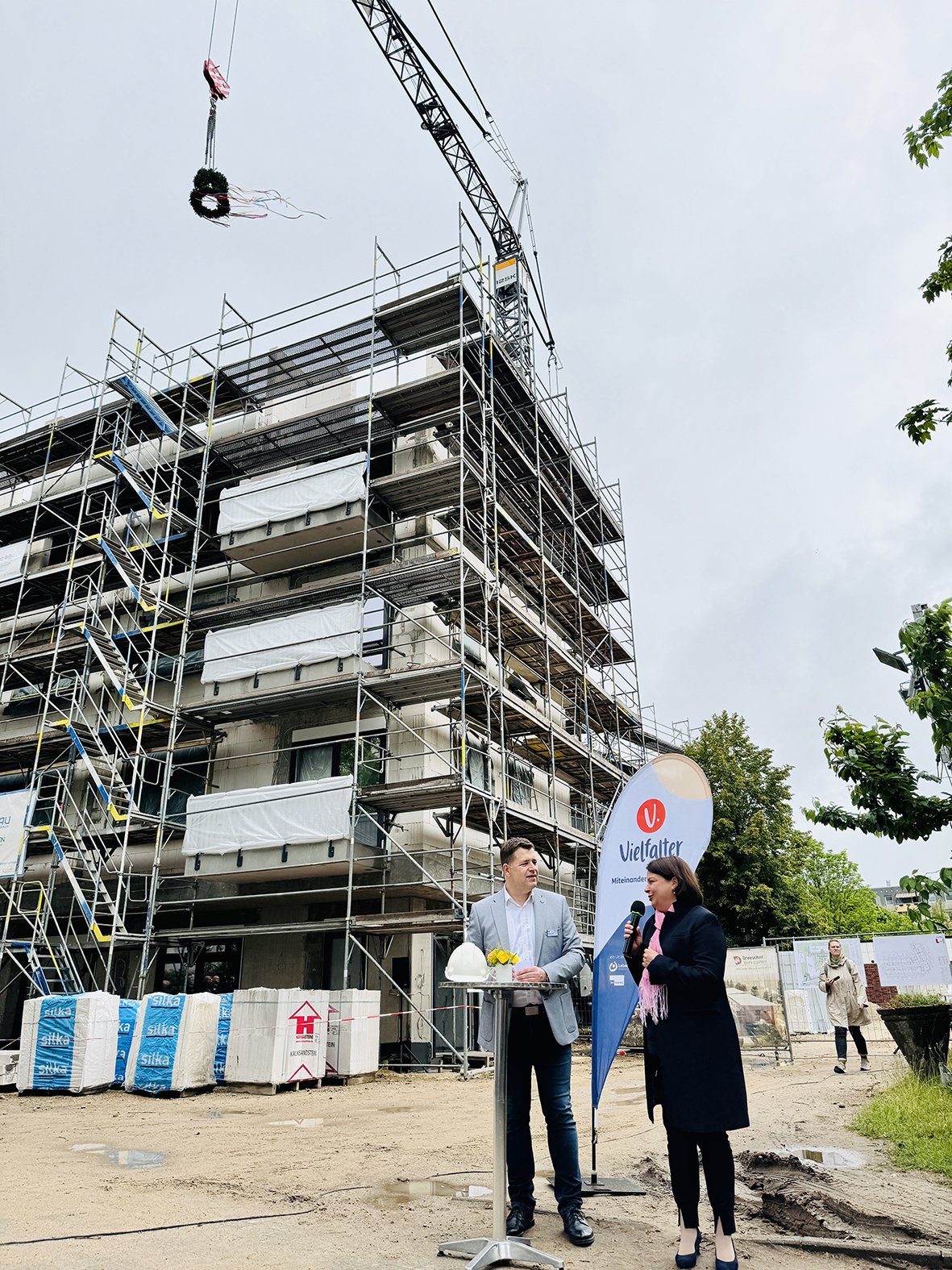 Ein Mann und eine Frau stehen beim Richtfest an einem Stehtisch vor einem mehrstöckigen Neubau