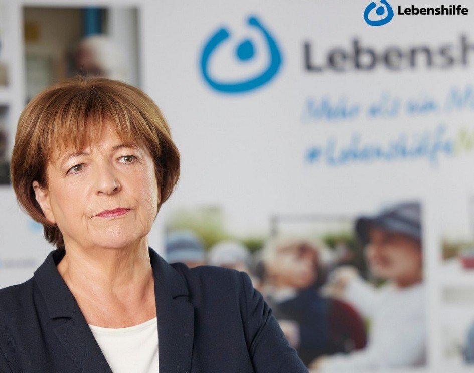 Eine Frau im Blazer steht vor einem Schild der Lebenshilfe