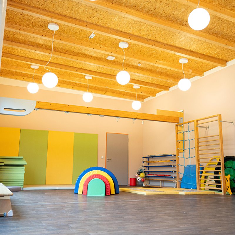 Ein großer, heller Turnraum der Kita mit hoher Holzdecke und runden Pendelleuchten. Im Raum verteilt stehen Klettergerüste, bunte Weichbodenmatten und Schaumstoff-Spielelemente.