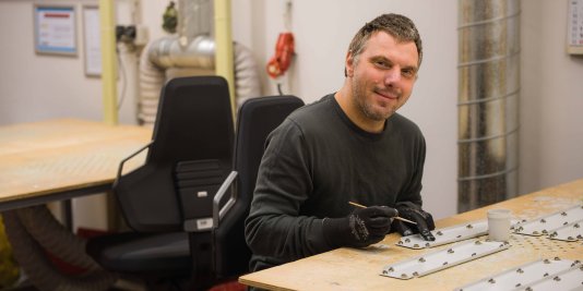 Ein Mann sitzt an seinem Arbeitsplatz in der Werkstatt und behandelt kleine Metallteile