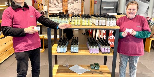 Ein Mann und eine Frau stehen neben einem Regal im Werkstattladen und präsentieren Seifen und Duschgels