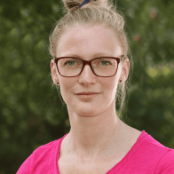 Nahaufnahme einer blonden Frau mit zusammengebundenen Haaren und einer roten Brille vor unscharfem grünen Hintergrund