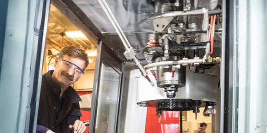 Ein Mann mit Schutzbrille und blauer Jacke arbeitet an einer Bohrmaschine