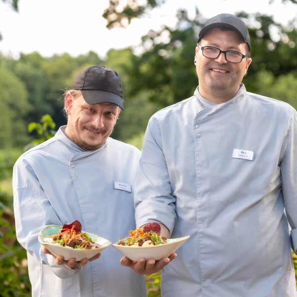 Zwei Mitarbeiter aus dem inklusiven Team des Bistros am Zoo. Sie halten Gerichte, die man dort essen kann.
