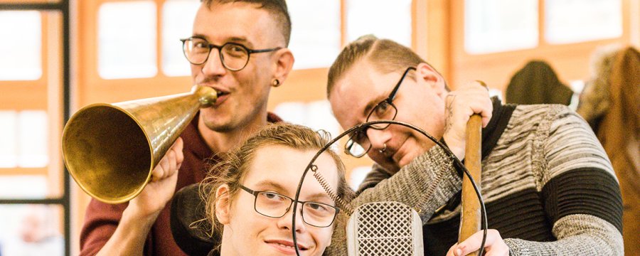 Drei Männer mit Brille stehen und sitzen hinter einem alten Mikrofon.