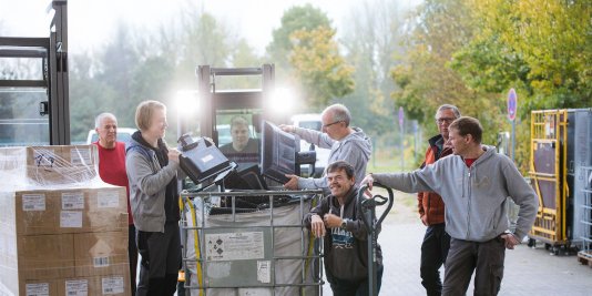 Eine Gruppe von Männern steht vor einem Gabelstapler und sortiert Elektroschrott in eine große Box.