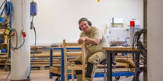 Mann mit Gehörschutz und Arbeitskleidung sitzt in der Holzwerkstatt und bearbeitet eine Stuhl