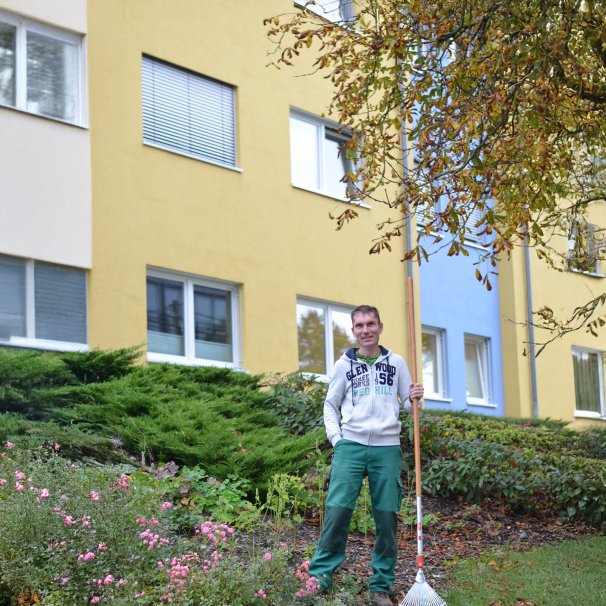 Mann in Arbeitskleidung steht mit einer Harke in der Grünanlage vor einem Neubaublock.