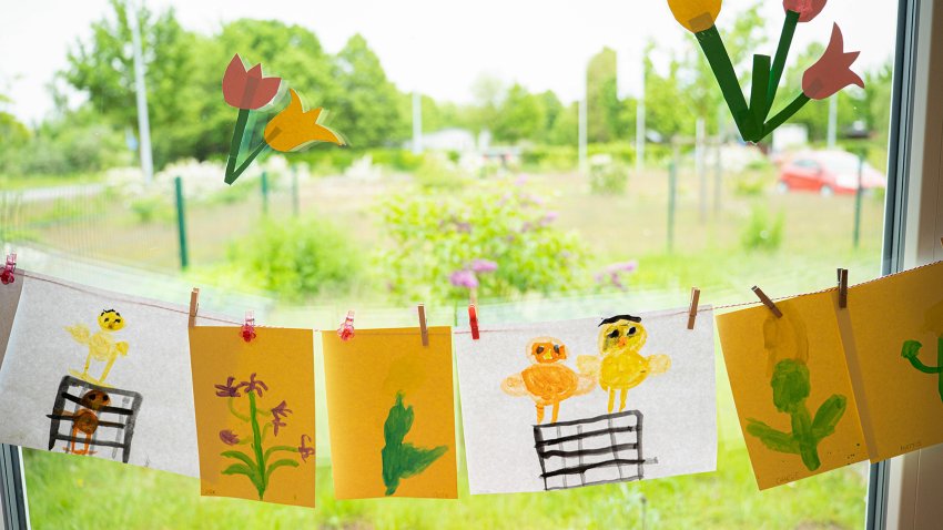 Bunte Kinderzeichnungen mit Küken- und Blumenmotiven hängen an einer Schnur vor einem Kita-Fenster. Auf der Scheibe kleben gebastelte Tulpen, dahinter ist eine Wiese sichtbar.