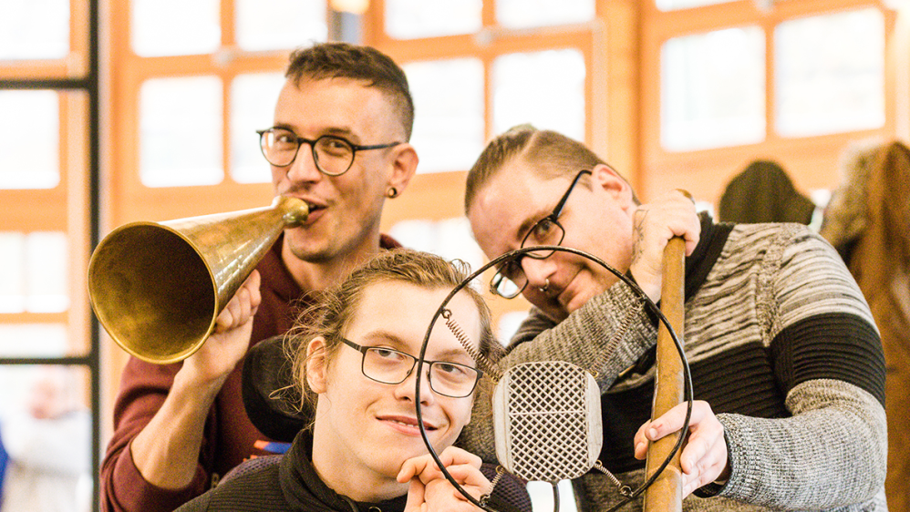 Drei Männer mit Brille stehen und sitzen hinter einem alten Mikrofon.