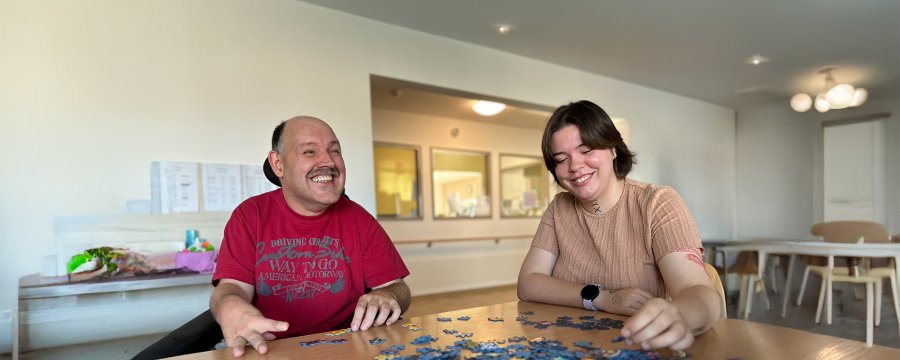 Ein Mensch mit Unterstützungsbedarf und eine junge Frau sitzen gemeinsam am Tisch und puzzeln.