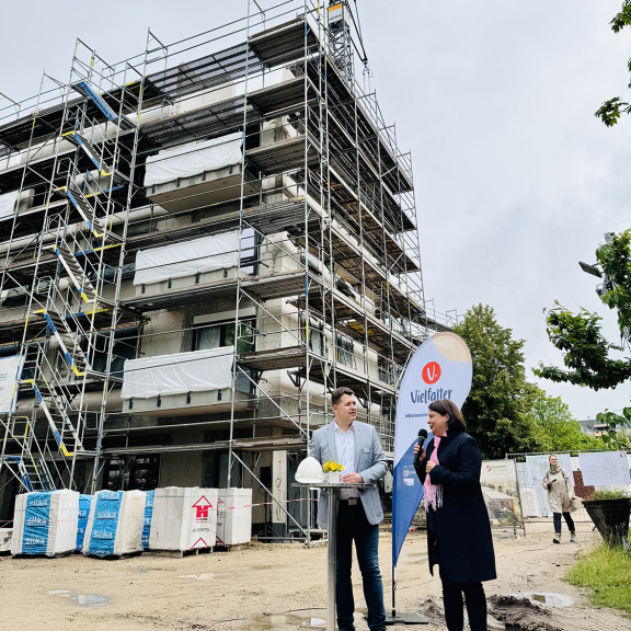 Ein Mann und eine Frau stehen beim Richtfest an einem Stehtisch vor einem mehrstöckigen Neubau