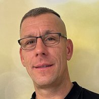 Nahaufnahme eine Mannes mit kurzen haaren und dunkler Brille vor unscharfen grünem Hintergrund