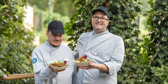 Zwei Mitarbeiter aus dem inklusiven Team des Bistros am Zoo. Sie halten Gerichte, die man dort essen kann.