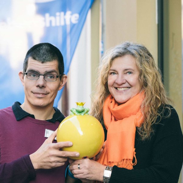 Ein Mann und eine Frau halten eine große gelbe Keramik mit einem Froschkönig darauf in ihren Händen.