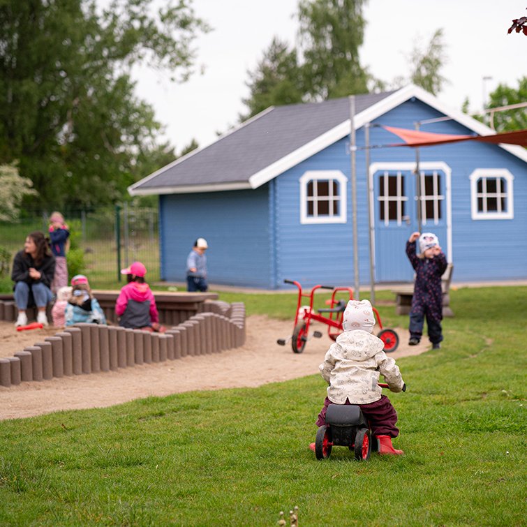 Ein Kleinkind fährt im Vordergrund auf einem Spielauto über die Wiese einer Kita. Dahinter spielen weitere Kinder im Sandkasten und im Garten vor einem blauen Holzhaus.