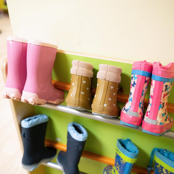 Mehrere Paare bunter Kindergummistiefel stehen ordentlich aufgereiht in einem Schuhregal der Kita. Die Stiefel zeigen verschiedene Muster wie Herzen und Schmetterlinge.