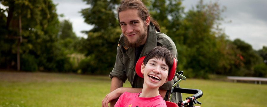 Ein junger Mann begleitet ein Kind im Rollstuhl durch den sommerlichen Park.