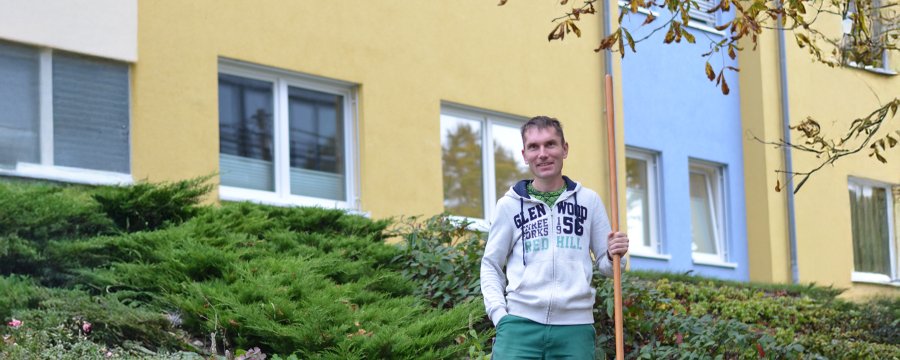 Mann in Arbeitskleidung steht mit einer Harke in der Grünanlage vor einem Neubaublock.