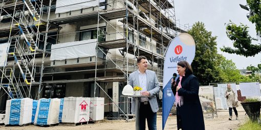 Ein Mann und eine Frau stehen beim Richtfest an einem Stehtisch vor einem mehrstöckigen Neubau