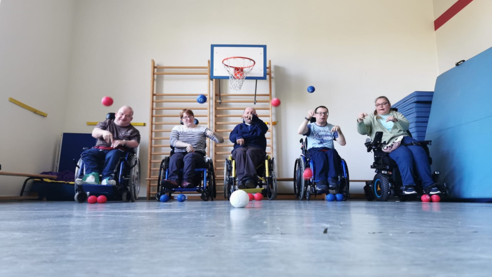 Fünf Rollstuhlfahrer trainieren in der Turnhalle das Boccia-Spiel.