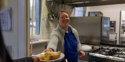 Junge Frau reicht lächelnd einen mit Essen gefüllten Teller über den Tresen